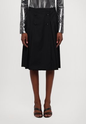 Mujer con falda plisada negra hasta la rodilla con botones, top plateado de manga larga y sandalias negras abiertas en fondo blanco.