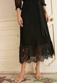 Jupe plissée noire avec un ourlet en dentelle festonnée, texture transparente et une taille à nouer assortie. Une paire de sandales à talons dorés est visible en bas.