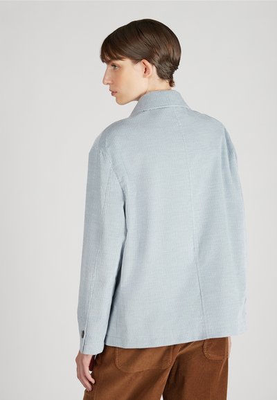 Jasnoniebieska, paskowana koszula sztruksowa z kołnierzykiem i guzicznymi mankietami, o luźnym kroju i widocznymi szwami, zestawiona z brązowymi spodniami sztruksowymi.