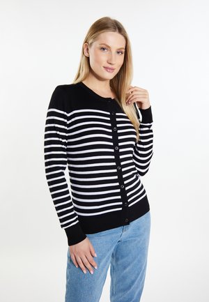 Vrouw met lang blond haar die een zwart-wit gestreepte, geknoopte cardigan en blauwe spijkerbroek draagt, staand tegen een witte achtergrond.