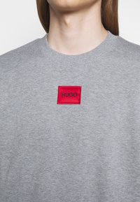 Gros plan sur une personne portant un sweat-shirt gris clair à col rond avec une petite étiquette carrée rouge portant l'inscription "HUGO" sur la poitrine.