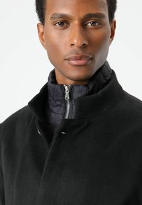 Zwarte wollen jas met hoge kraag, voorzien van een getextureerde zwarte binnenlaag en een zilveren ritssluiting. Gladde stof met een gestructureerd ontwerp.