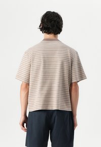 T-shirt à manches courtes, col rond, en beige avec des rayures horizontales marron clair. Tissu doux, coupe décontractée et ourlet court.