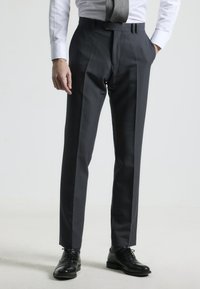Pantalones de vestir en gris carbón, con un frente plano, trabillas para cinturón y dos bolsillos laterales, combinados con una camisa blanca y una corbata gris. Zapatos negros.