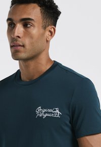 Donker turquoise T-shirt met ronde hals, voorzien van een witte geborduurde "Original Penguin" logo op de linkerkant van de borst. Zachte, gladde stof.
