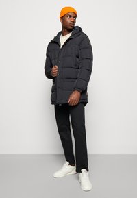 Giacca a vento nera con cappuccio, tasche frontali e chiusure a pressione. Il modello indossa un berretto arancione, pantaloni neri e sneaker bianche.