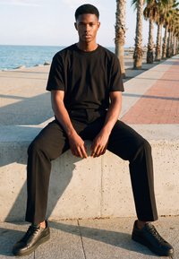Jeune homme en tenue noire assis sur un rebord en béton près de la promenade en bord de mer bordée de palmiers au coucher du soleil.