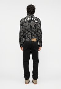 Μπουφάν από μαύρο τζιν με υφή σχεδίου, λευκά γράμματα "ICECREAM" στην πλάτη και καφέ ετικέτα στη μέση. Φορεμένο με μαύρο παντελόνι.