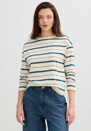 Mujer joven con camiseta de manga larga color crema con rayas horizontales azules y vaqueros azul sueltos de talle alto sobre fondo blanco.