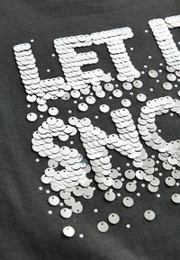 Grå stof med sølvfarvede pailletter, som danner teksten "LET IT SHINE", spredte pailletter omkring bogstaverne, og en glat, reflekterende overflade.