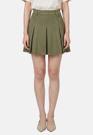 Donna che indossa una gonna a pieghe verde oliva, un top bianco e décolleté beige con punta a punta, in piedi con le braccia rilassate ai lati.