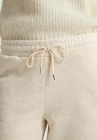 Pantalons de survêtement beige avec taille élastique et cordon de serrage. Tissu lisse avec une texture subtile; embouts métalliques sur le cordon.