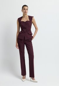 Gilet bordeaux su misura con scollatura quadrata, quattro bottoni e tasche frontali, abbinato a pantaloni eleganti coordinati e tacchi alti beige.