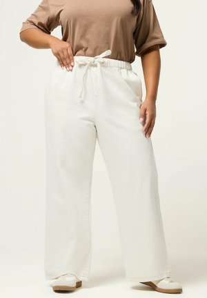 Kvinde iført løse off-white bukser med snøre, beige kortærmet top og hvide slip-on sko, stående med den ene hånd på hoften.