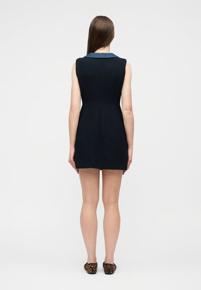 Γυναίκα που στέκεται με την πλάτη, φορώντας ένα μαύρο αμάνικο φόρεμα με μπλε γιακά και φλατ παπούτσια με λεοπάρ σχέδιο.