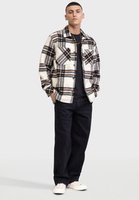 Veste écossaise en flanelle crème, noire et orange avec deux poches avant, portée avec un pantalon noir et des baskets blanches.