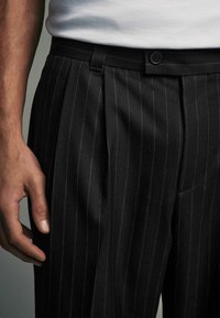 Black pinstriped trousers made of fabric with a smooth texture. Features pleats, a button closure, and belt loops. Hand resting on the fabric.