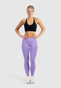 Zwarte sportbeha met geplooid detail, gecombineerd met effen lavendel leggings. Model staat zelfverzekerd met handen op de heupen, en draagt witte sneakers.
