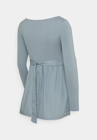 Top s dlouhým rukávem z měkké světle modré tkaniny, má přiléhavý živůtek a rozšířený spodní lem s mašlí vzadu. Hladká textura.