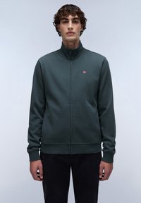 Sudadera de cremallera en verde oscuro con cuello alto, puños y dobladillo de canalé. Presenta un pequeño logo de bandera en el pecho y una textura de tela suave.