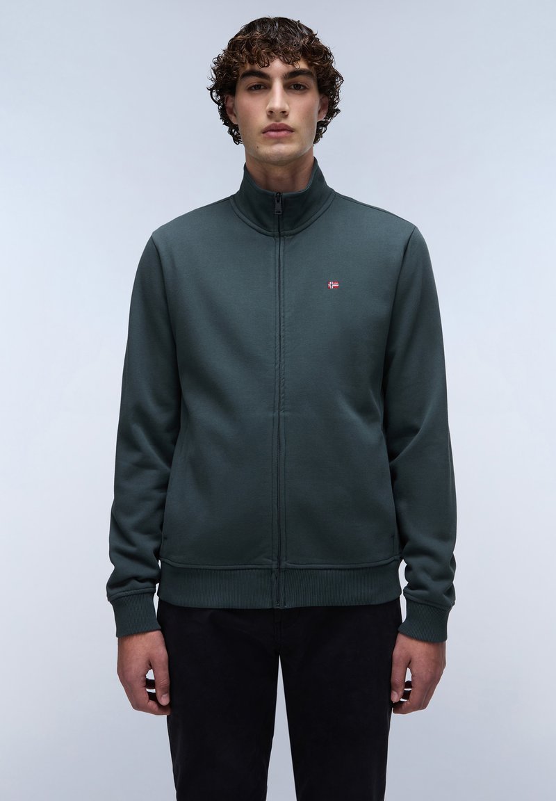 Sudadera de cremallera en verde oscuro con cuello alto, puños y dobladillo de canalé. Presenta un pequeño logo de bandera en el pecho y una textura de tela suave.