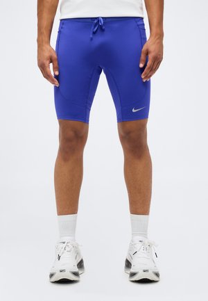 Mężczyzna w niebieskich dopasowanych sportowych krótkich spodenkach z logo Nike, białych skarpetkach i białych butach do biegania stoi na jednolitym tle.