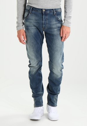 Mężczyzna w wyblakłych niebieskich dżinsach, szarej długorękawowej koszuli i białych tenisówkach, stojący z rękami swobodnie opuszczonymi wzdłuż ciała.