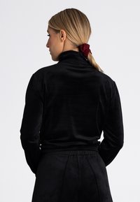 Czarny welurowy top z wysokim kołnierzem i długimi rękawami, o gładkiej fakturze. Modelka pokazuje widok z tyłu i ma na sobie czerwoną gumkę do włosów.