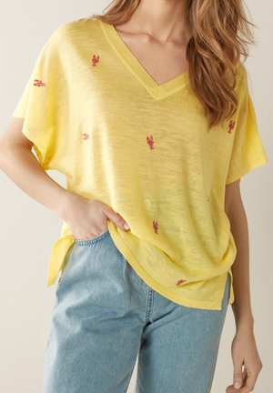 Femme portant une chemise jaune ample à col en V avec de petites imprimés homards rouges, associée à un jean taille haute bleu clair.