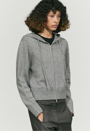 Jeune femme portant un sweat à capuche gris clair zippé avec capuche et poignets côtelés, mains dans les poches d’un pantalon gris foncé, debout devant un fond uni.