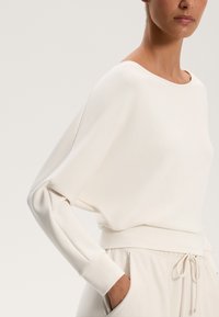 Cămașă albă cu mâneci lungi, confecționată dintr-un material moale și neted, cu un decolteu larg și o croială lejeră. Dispune de un brâu ajustat care completează partea de jos în culori deschise.