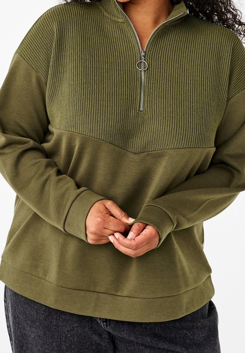 Personne portant un pull vert olive avec une section supérieure côtelée et une fermeture éclair à anneau argenté, mains doucement jointes devant.