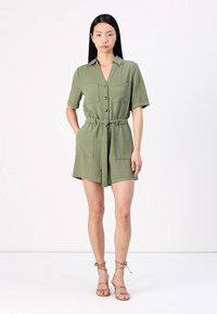 Combinaison utilitaire verte à manches courtes avec col, devant à boutons, deux poches poitrine et taille nouée. Présente un tissu texturé et une coupe décontractée.