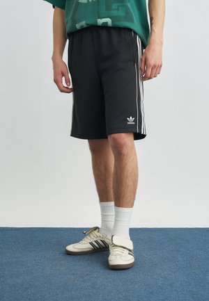 Osoba oblečená v čiernych šortkách Adidas s bielymi pruhmi a logom, biele ponožky do členkov, a biele tenisky Adidas na modrom koberci.