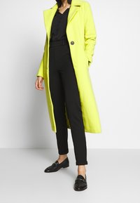 Pitkä neonkeltaisen takki eiolkaistulla kauluksella ja yhdellä mustalla napilla, päällä musta yläosa ja mustat housut, yhdistetty mustiin loafer-kenkiin.