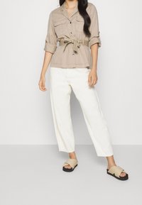 Beige jakke med bælte og brystlommer, kombineret med hvide bukser med vide ben og taupe sandaler med et krydsremdesign.