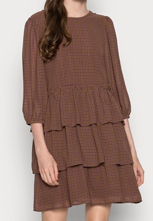 Vrouw draagt een bruin-paars geruite jurk met meerdere lagen en driekwart mouwen tegen een effen lichte achtergrond.