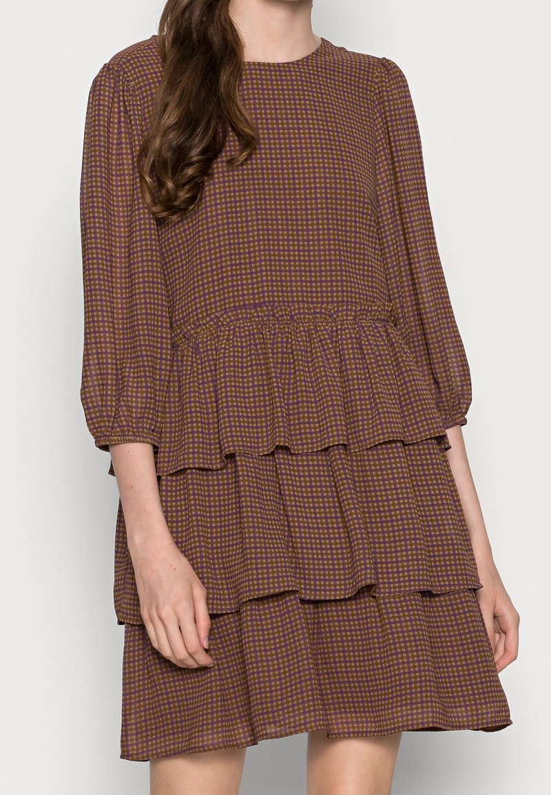 Femme portant une robe à étages écossaise marron et violette avec des manches trois-quarts, sur un fond clair uni.