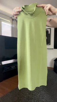 Pessoa segurando um vestido sem mangas de gola alta verde limão dentro de uma sala de estar com uma televisão e fotos emolduradas ao fundo.