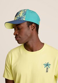 La casquette a un corps turquoise avec une visière bleue et un motif de palmier sur le devant. Le t-shirt est jaune avec un logo de palmier brodé.