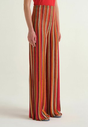 Personne portant un pantalon taille haute, à jambes larges, avec des rayures verticales dans des tons rouges, orange, jaune, rose et marron, assorti à des sandales bleues.