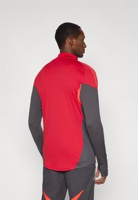 Rød og grå langærmet atletisk trøje med høj krave og farveblokdesign med kontrasterende orange accenter. Glat tekstur, kropsnær pasform.
