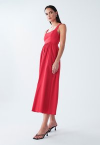Rotes ärmelloses Maxikleid mit quadratischem Ausschnitt, geraffter Taille und glatter Textur; getragen mit schwarzen Sandaletten mit Absatz.