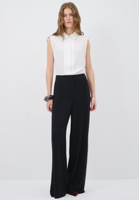 Witte mouwloze blouse met een knoopsluiting aan de voorkant, gecombineerd met zwarte wijde broekspijpen. Een doorzichtige armband siert de arm.