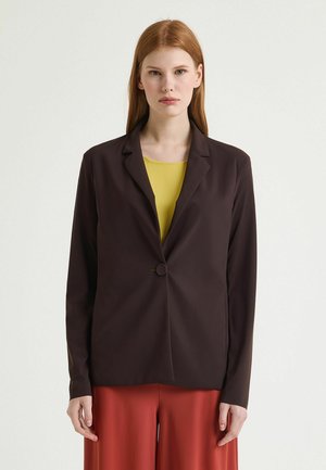 Frau mit roten Haaren, die einen dunkelbraunen Ein-Knopf-Blazer über einem gelben Oberteil und roten, weit geschnittenen Hosen trägt, vor einem schlichten Hintergrund stehend.