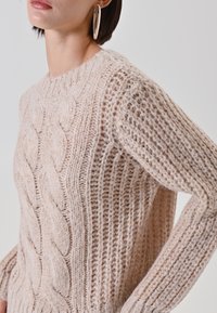 Maglione lavorato a maglia in morbido filato beige con motivo a trecce, trama a coste e scollo tondo. Dettaglio sulle maniche che mostra una vestibilità leggermente ampia.