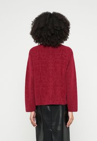 Κόκκινο πουλόβερ με απαλή υφή και διακριτικό σχέδιο, με μακριά μανίκια και ψηλό γιακά, συνδυασμένο με κομψή μαύρη δερμάτινη φούστα.