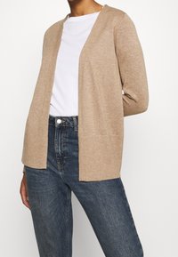 Beige cardigan made of soft fabric with an open front, featuring long sleeves and a relaxed fit. Paired with blue jeans and a white shirt.