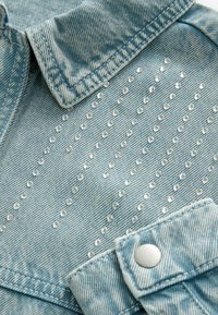 Cămașă din denim de culoare albastru deschis, cu guler, dotată cu nituri argintii dispuse vertical pe față și cu manșete cu butoni. Material texturat.