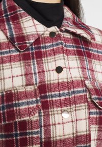 Koszula flanelowa w czerwono-białą kratę z zapięciem na guziki, kołnierzem oraz kieszenią na piersi z metalicznym akcentem. Miękka w dotyku.